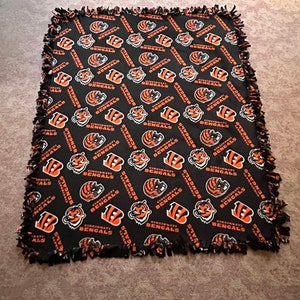 May include: A black fleece blanket with a repeating pattern of orange and white Cincinnati Bengals logos. The blanket has a knotted fringe border.