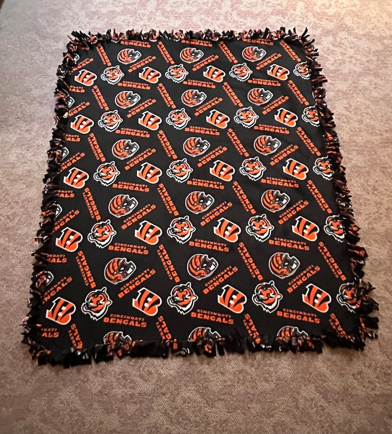May include: A black fleece blanket with a repeating pattern of orange and white Cincinnati Bengals logos. The blanket has a knotted fringe border.