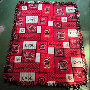 May include: Red, white, and gray fleece blanket with a University of South Carolina logo and Gamecocks text. The blanket has a knotted fringe border.