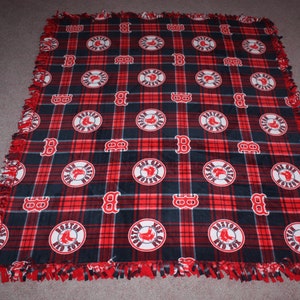 May include: Red and black fleece blanket with a plaid pattern and a repeated design of white circles with the words "Boston Red Sox" inside. The blanket has a frayed edge.