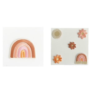May include: A set of five embroidered iron-on patches featuring a rainbow, a sun, and three flowers. The rainbow is in shades of brown, pink, and orange. The sun is gold with a white outline. The flowers are in shades of brown, orange, and pink.