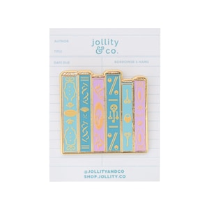 May include: A gold-edged enamel pin featuring a stack of stylized books in shades of teal, lavender, and gold. Each book has unique decorative elements. The pin is set against a library card background with the text "jollity & co."