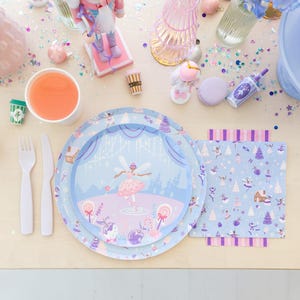 May include: A festive table setting with a light blue and pink theme. Includes plates with a ballerina design, a matching napkin, and a cup of orange juice. Decorative elements include a nutcracker, ornaments, and a small coffee cup.