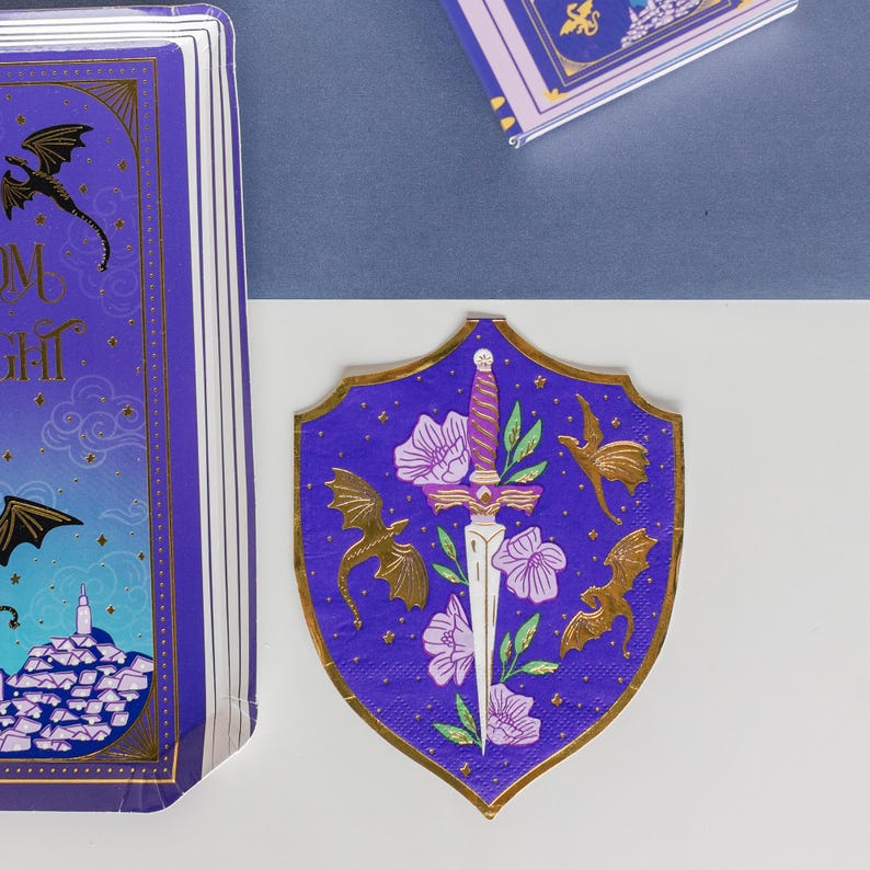 May include: Purple shield-shaped paper napkin with gold trim, featuring a sword, flowers, and gold dragon illustrations. The napkin is part of a set with a matching book and other items, all with a fantasy theme.