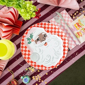 Op de afbeelding: Een feestelijke kersttafelsetting met een kerstmanbord, een rood-wit geblokte servet en een servet met de tekst "Merry Christmas". De tafel is versierd met ornamenten, bloemen en confetti.