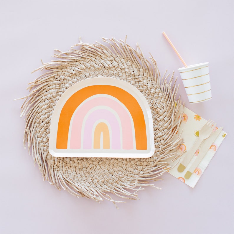 May include: A rainbow-themed paper plate with a woven straw placemat, a paper cup with gold stripes, and a set of wooden forks with a floral patterned napkin.