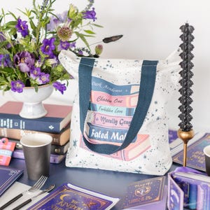 Op de afbeelding: Een witte draagtas met marineblauwe handvatten, met een stapel boektitels zoals "Dark Academia" en "Fated Mate". De tas heeft een spat- en sterrenontwerp. Een vaas met paarse bloemen en een zwarte kaars zijn ook zichtbaar.