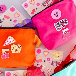 May include: Two bright pink and orange zippered pouches with various colorful patches and embroidery. The patches include hearts, cherries, pretzels, pizza slices, and smiley faces. The pouches have the word "Love" embroidered on them.