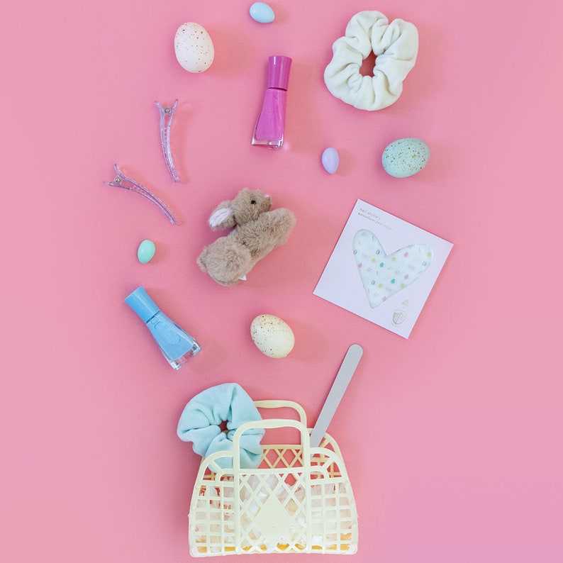 May include: A yellow plastic tote bag with a blue scrunchie, a brown bunny, nail polish, and pastel Easter eggs on a pink background.