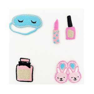 Puede incluir: Un conjunto de cinco parches bordados para planchar que incluyen una máscara para dormir, pintalabios, esmalte de uñas, un frasco de perfume y zapatillas de conejo. Los parches son de color rosa, azul y negro.