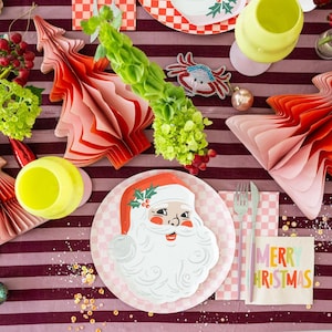 Op de afbeelding: Een feestelijke kersttafelsetting met een kerstmanbord, rode en roze papieren boomversieringen en een servet met "MERRY CHRISTMAS". De tafel is gedekt op een bordeaux gestreept tafelkleed.