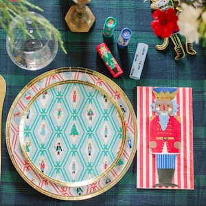 Puede incluir: Una mesa festiva navideña con platos estampados, una servilleta de cascanueces y tubos decorativos. Los platos tienen un borde dorado y un diseño geométrico turquesa y blanco con figuras navideñas. La servilleta presenta un cascanueces.