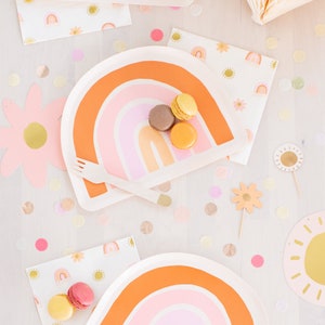 May include: Two rainbow-themed paper plates with a pink, orange and yellow design. The plates are decorated with a rainbow arch and are surrounded by confetti and other party decorations.