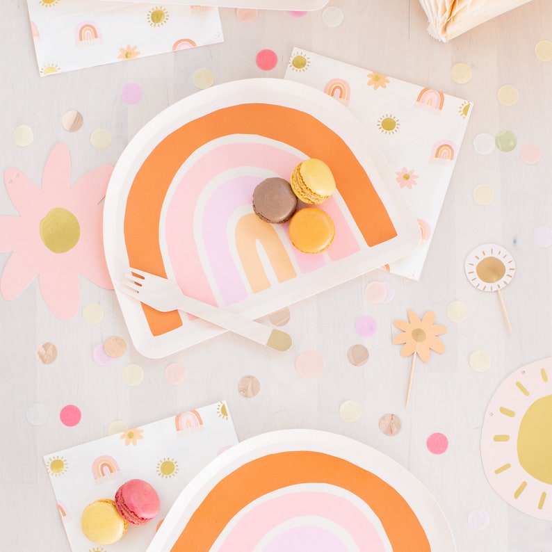 May include: Two rainbow-themed paper plates with a pink, orange and yellow design. The plates are decorated with a rainbow arch and are surrounded by confetti and other party decorations.