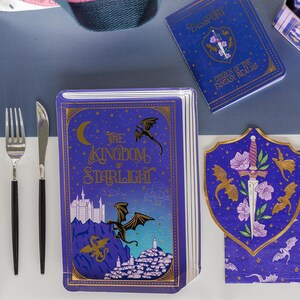 May include: A fantasy-themed party set featuring a book-shaped plate with the text "The Kingdom of Starlight", a passport, and a shield-shaped napkin. The set is decorated with dragons, castles, and floral accents in gold and purple.