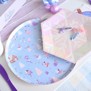 Op de afbeelding: Een lichtblauwe ronde papieren bord met een grillig balletthema-ontwerp, met ballerina's, peperkoekhuisjes en snoep. Een zeshoekig bord met sneeuwvlokkenpatroon en twee decoratieve haarclips zijn ook zichtbaar.