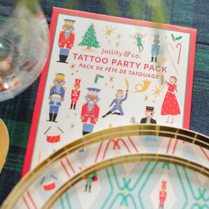 Può includere: Un pacchetto di tatuaggi festivo con illustrazioni di schiaccianoci, un albero di Natale e altri personaggi a tema natalizio. Il pacchetto è etichettato "jollity & co. TATTOO PARTY PACK" con la traduzione francese "PACK DE FÊTE DE TATOUAGE".