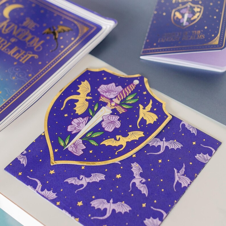May include: A collection of fantasy-themed stationery items. The items include a notebook, a shield-shaped card, and a patterned card, all in shades of blue and gold. The shield card features a sword, flowers, and dragon illustrations. The notebook has the text "The Kingdom of Twilight".