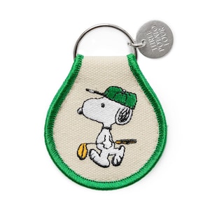 Può includere: Un portachiavi di Snoopy con un design in tela bianco sporco bordato di verde. L'immagine ricamata raffigura Snoopy con un berretto da golf verde, che tiene una mazza da golf e una sacca da golf gialla. È attaccato un piccolo tag in metallo con la scritta "THREE POTATO FOUR".