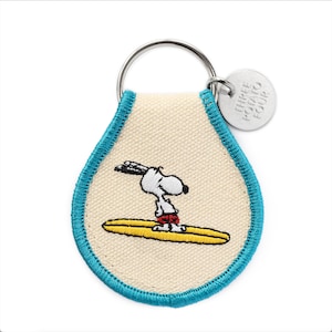 Op de afbeelding: Een sleutelhanger van wit canvas met een blauwe rand met een geborduurd beeld van Snoopy die surft. De sleutelhanger heeft een zilveren metalen ring en een zilveren metalen label met de tekst "Three Potato Four".