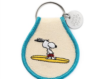 Könnte beinhalten: Ein Schlüsselanhänger aus weißem Canvas mit blauem Rand, der ein gesticktes Bild von Snoopy beim Surfen zeigt. Der Schlüsselanhänger hat einen silbernen Metallring und ein silbernes Metallschild mit der Aufschrift "Three Potato Four".