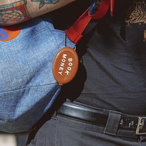 Op de afbeelding: Een bruine, ovale portemonnee met de witte woorden "BOOK MONEY". Het is bevestigd aan een blauwe denim tas met een rode band. De afbeelding is een close-up.