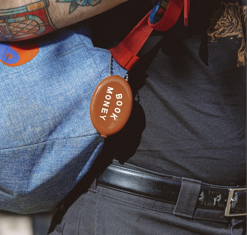 Op de afbeelding: Een bruine, ovale portemonnee met de witte woorden "BOOK MONEY". Het is bevestigd aan een blauwe denim tas met een rode band. De afbeelding is een close-up.