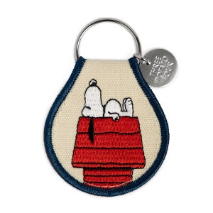 May include: A white canvas keychain with a blue border featuring an embroidered image of Snoopy lying on top of his red doghouse. The keychain has a silver metal ring and a silver tag that reads "Three Potato Four".