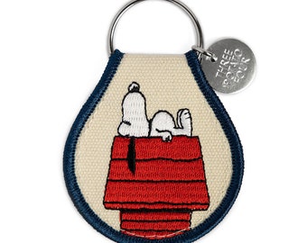 May include: A white canvas keychain with a blue border featuring an embroidered image of Snoopy lying on top of his red doghouse. The keychain has a silver metal ring and a silver tag that reads "Three Potato Four".
