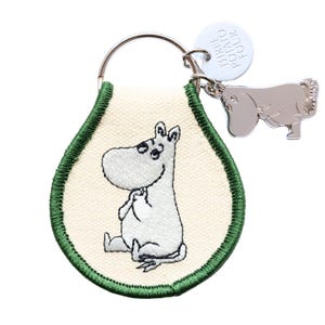 May include: A keychain featuring a white embroidered character on a cream-colored background with a green border. A silver charm and a tag with the text "THREE POTATO FOUR" are attached to the keyring.