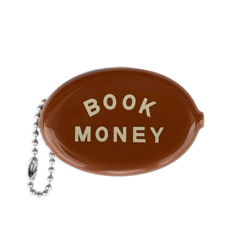Op de afbeelding: Een bruin, ovaalvormig muntzakje met de witte tekst "BOOK MONEY". Het zakje heeft een glanzende afwerking en een zilverkleurige ketting. Handig voor het bewaren van kleingeld.