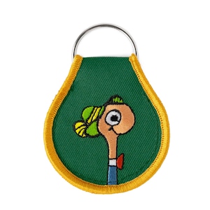 May include: Green fabric keyring with a yellow border. The keyring features an embroidered cartoon character with a large eye, a green hat, and a red bow tie.
