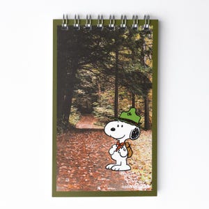 May include: Spiral-bound notepad featuring a forest scene with a path and Snoopy in a green hat and backpack. The notepad has a green border and a metal spiral binding at the top. The image includes the text "© 2023 Peanuts".