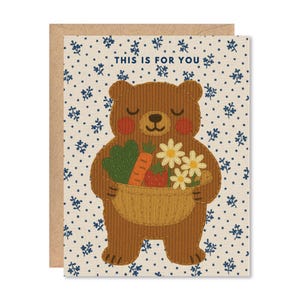 May include: A greeting card featuring a brown bear holding a basket of vegetables and flowers. The bear has rosy cheeks and a smiling expression. The card has the text "THIS IS FOR YOU" and a floral pattern on a cream background.