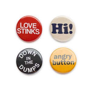 May include: Four round button badges with different sayings. The first badge is red with white text that says "LOVE STINKS". The second badge is beige with blue text that says "Hi!". The third badge is black with white text that says "DOWN IN THE DUMPS". The fourth badge is yellow with black text that says "angry button".