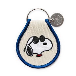 Pode incluir: Um chaveiro Snoopy com fundo branco e borda azul. O chaveiro apresenta um design Snoopy bordado com óculos de sol pretos e uma gola vermelha. Uma pequena etiqueta de metal diz "THREE POTATO FOUR".
