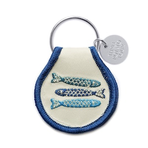 May include: A white fabric keychain with a blue border and three embroidered blue fish. The keychain has a silver ring and a silver tag that says "Three Potato Four".