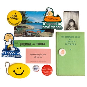 K&ouml;nnte beinhalten: Eine Sammlung von Vintage-Ansteckern und Kn&ouml;pfen mit Peanuts-Charakteren, einem Smiley und einem Herzen. Au&szlig;erdem gibt es Postkarten, ein Buch mit dem Titel "The Observer's Book of Garden Flowers" und ein kleines Schild mit der Aufschrift "Special for Today".