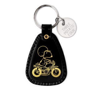 May include: Black leather keychain featuring a gold Snoopy illustration riding a motorcycle. The keychain includes a silver ring and a small, round tag with the text "THREE POTATO FOUR". The design is from ©2025 PNTS.