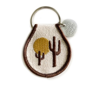 May include: A brown and white key chain with a brown embroidered cactus and a yellow sun. The key chain has a silver tag that says "Three Potato Four".