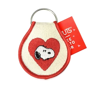May include: A Snoopy keychain featuring a red heart with Snoopy inside. The keychain has a white background with a red border and a silver key ring. A red tag with white text is attached.