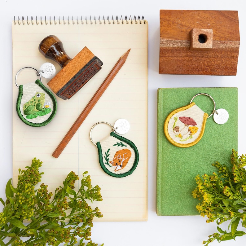 May include: An overhead shot features a collection of nature-themed keychains, a wooden stamp, and a pencil on a lined notepad. The keychains depict a frog, a fox, and mushrooms. Green and yellow foliage accents the scene.