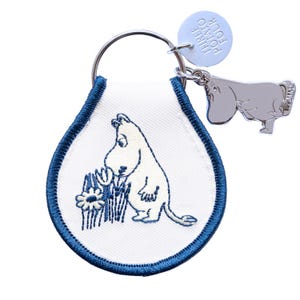 May include: A white keychain with a blue embroidered border and a cartoon character design. The keychain features a silver metal ring, a round tag with text, and a silver hippo charm. The cartoon character is white with a black outline.