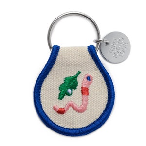 May include: A blue and white key chain with a small embroidered worm and leaf design. The key chain has a metal tag that says "Three Potato Four".
