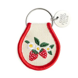 May include: A cream-colored embroidered keychain with a red border, featuring strawberries and white flowers. The keychain has a silver ring and a small tag that reads "THREE POTATO FOUR."