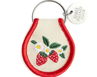 Puede incluir: Llavero bordado de color crema con borde rojo, con fresas y flores blancas. Tiene un anillo plateado y una etiqueta que dice "THREE POTATO FOUR".
