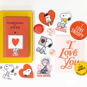 May include: A collection of Snoopy-themed stickers, buttons, and a book. The stickers feature Snoopy in various poses, hearts, and phrases like "I Love You" and "I Love Snoopy." The book is titled "The Observer's Book of Dogs."