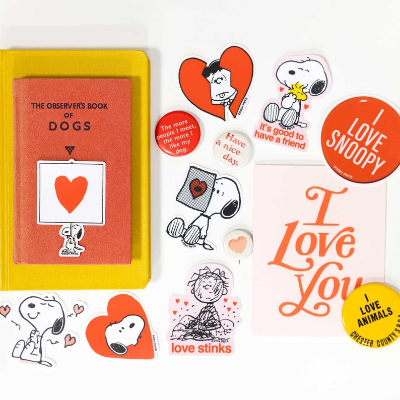 May include: A collection of Snoopy-themed stickers, buttons, and a book. The stickers feature Snoopy in various poses, hearts, and phrases like "I Love You" and "I Love Snoopy." The book is titled "The Observer's Book of Dogs."
