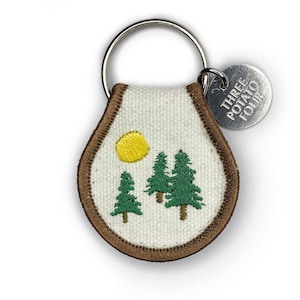 May include: A white canvas keychain with a brown border and an embroidered design of three green trees and a yellow sun. The keychain has a silver metal ring and a silver tag that reads "Three Potato Four".