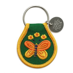 May include: A green and yellow fabric keychain with a butterfly and flowers embroidered on it. The keychain has a metal ring and a metal tag that says "THREE POTATO FOUR".